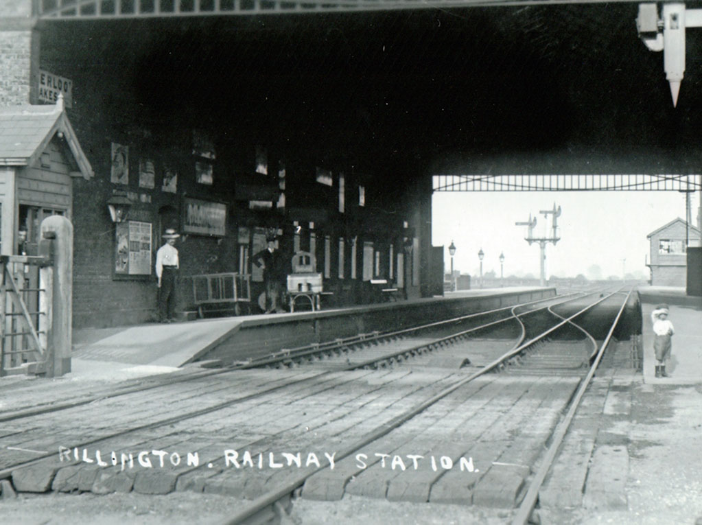 Rillington Railway Station beneath the overall roof, with passengers, a little boy, luggage on a porter's trolley, advertisements, tracks and signals.